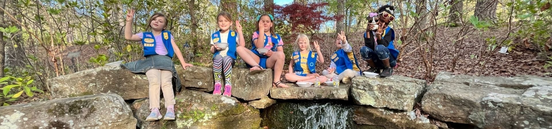  six daisy girl scouts making the girl scout sign and sitting on rocks on top of a water feature 