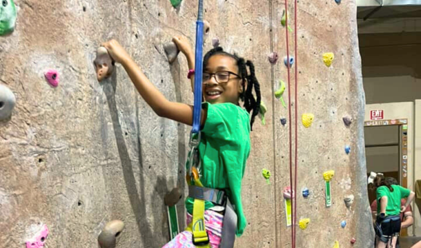 Girl in a green t-shirt climbing a rock climbing wall and smiling at the camera