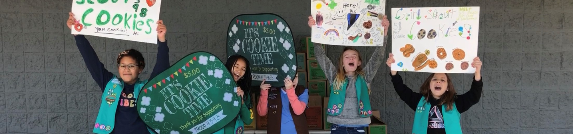  Five girls hold up cookie signs at an outdoor cookie booth 