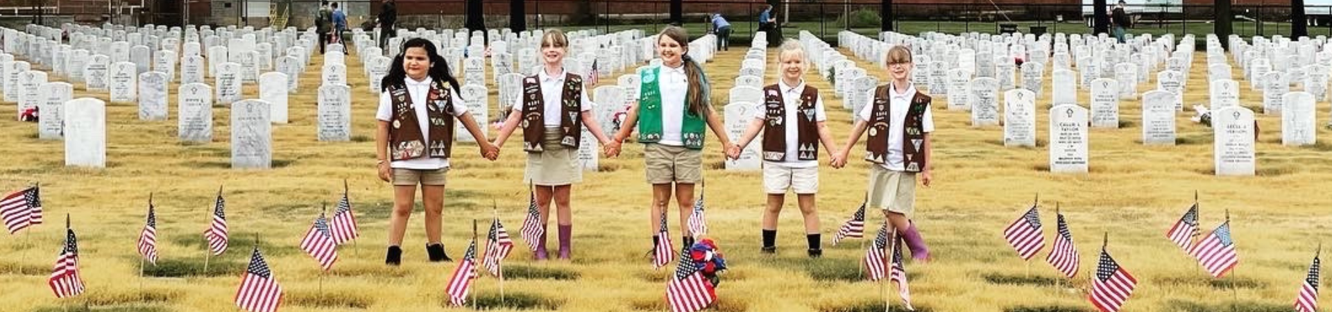  group of 5 girl scouts hold hands at a cemetery where they have placed flags 