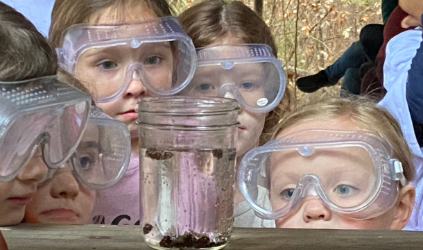 extreme close up of five young girl scouts wearing safety glasses and looking a jar with clear liquid and small brown objects