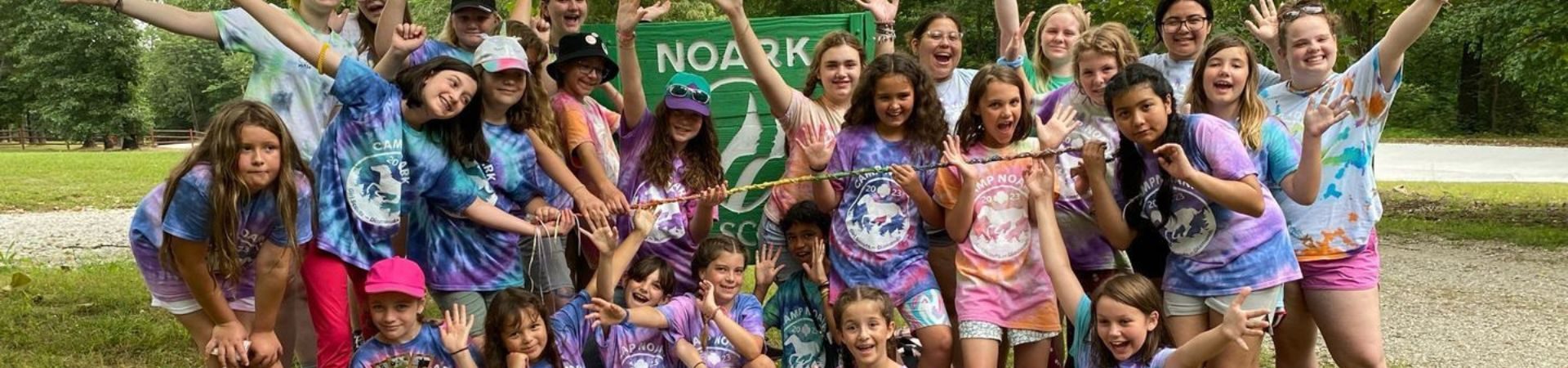  large group of girls in tie-dye summer camp t-shirts posed in front of Camp NOARK sign 
